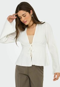 Haut en jersey blanc à côtes avec de longues manches évasées et une fermeture à nouer sur le devant. Il présente un col en V et un ourlet évasé. Porté par-dessus une couche intérieure beige.