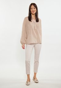 Blusa lunga beige chiaro, testurizzata, con bottoni, abbinata a pantaloni fitted beige chiaro e ballerine beige con un accento decorativo.