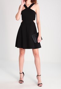 Svart ärmlös klänning med korsade axelband och en tajt överdel, som vidgar sig till en skaterkjol. Matchas med en svart kuvertväska och remsandaler.