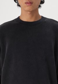 Črna crewneck majica iz mehkega blaga, ki ima rebrast ovratnik z gladko teksturo in minimalističen dizajn brez vidnih logotipov.