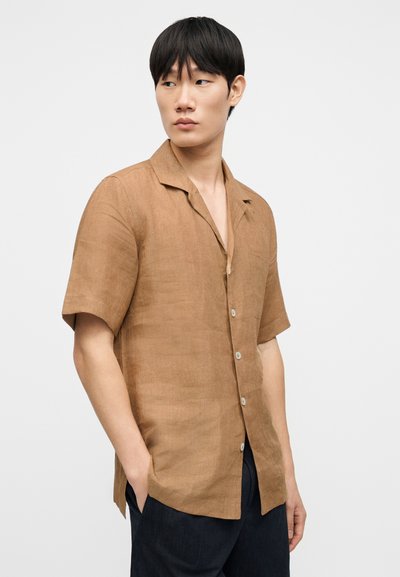Chemise marron en lin à manches courtes avec col campagnard et fermeture à boutons, présentant une texture lisse et des fentes latérales pour le confort.