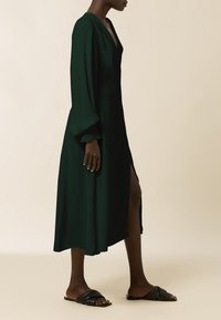 Largo vestido verde oscuro hecho de tela suave, con escote en V, mangas abullonadas y una abertura lateral. Combinado con sandalias deslizantes negras.