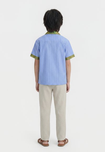 Άτομο όρθιο, γυρισμένο πίσω, φοράει μπλε πουκάμισο με κοντό μανίκι και πράσινες λεπτομέρειες, μπεζ παντελόνι και καφέ σανδάλια σε λευκό φόντο.