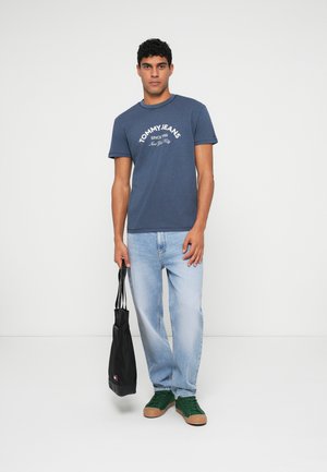 Menns marineblå bomull t-skjorte med "TOMMY JEANS" logo, sammen med lys blå jeans og mørke grønne sneakers. Holder en svart tote bag.