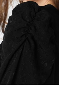 Épaule de femme avec tissu noir à manches ballons présentant des motifs subtils texturés d'étoiles et de crânes.