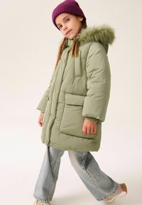 Grüne Puffjacke mit einem Kapuze aus Kunstfell, vorderen Taschen und elastischen Bündchen, kombiniert mit hellblauen weiter geschnittenen Jeans und einer lila Mütze.