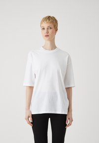 Tricou alb supradimensionat din bumbac, cu decolteu rotund și mâneci scurte. Textură netedă, tiv drept, purtat cu pantaloni negri.