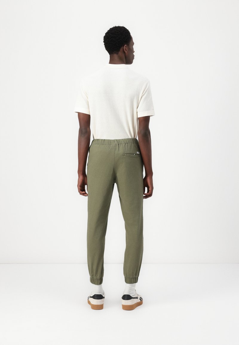 Pantalones joggers verde oliva con cintura y puños elásticos, material de mezcla de algodón, que presentan un bolsillo trasero y se combinan con una camiseta lisa color crema.