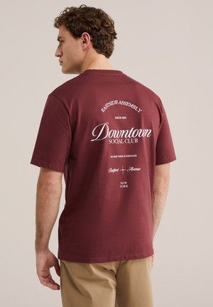 Bărbat cu păr creț purtând un tricou bordo cu text alb „Downtown Social Club” și pantaloni bej, văzut din spate pe un fundal simplu.