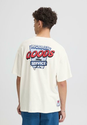 Krēmkrāsas kokvilnas T-krekls ar lielu grafiku uz muguras un sarkanu un zilu tekstu "WORKER GOODS SERVICE." Īsas piedurknes, brīvs piegriezums.