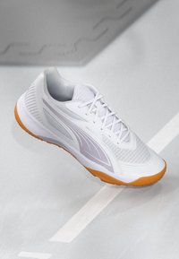 Białe buty sportowe z jasnoszarymi akcentami i pomarańczową podeszwą leżące na jasnoszarym podłożu z delikatnymi liniami oznaczeń.