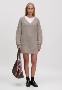 Hellgraues, gestricktes Kleid mit V-Ausschnitt, langen Ärmeln und strukturiertem Finish. Kombiniert mit einer braunen Tasche, die mit einem rosa karierten Schal verziert ist.