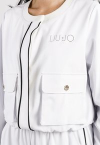 Giacca bianca con chiusura a zip, piping nero, due tasche anteriori e dettaglio del logo in strass. Realizzata in tessuto liscio e leggero.
