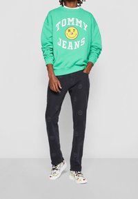 Green sweatshirt with "TOMMY JEANS" and smiley face graphic, paired with black pants featuring smiley patterns, and colorful, patterned sneakers.