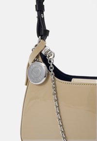 Beige lakatun nahan käsilaukku, jossa on kaareva muoto, musta nahkahihna, hopeinen ketjuyksityiskohta ja pyöreä logokoriste kiinnitettynä.