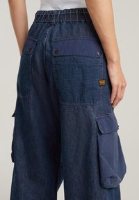 Siniset farkut, joissa on leveät lahkeet, joustava vyötärö, kaksi takataskua ja suuret sivutaskut. Teksturoitu kangas, jossa on kontrastivärejä.