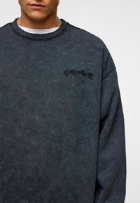 Donkergrijze sweatshirt met een gewassen textuur, voorzien van een ronde halslijn en geborduurd logo-opdruk op de borst. Vrije pasvorm.