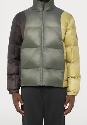 Homme portant une doudoune bicolore avec des sections olive, marron foncé et jaune, zippée, et un pantalon noir.