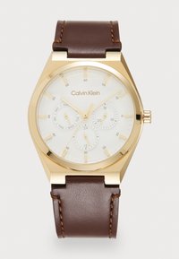 Reloj cronógrafo de tono dorado con una esfera blanca texturizada, correa de cuero marrón y acentos cosidos. Presenta la fecha y el día de la semana.