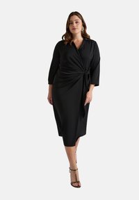 Robe noire midi avec un design croisé, manches longues et un détail en nœud à la taille. Tissu lisse avec une texture élégante, associé à des sandales à lanières.