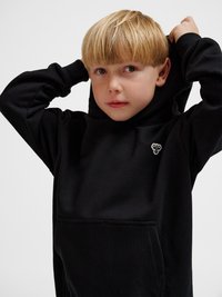 Jonge jongen met blond haar die een zwarte hoodie draagt en de capuchon over zijn hoofd trekt tegen een effen lichte achtergrond.