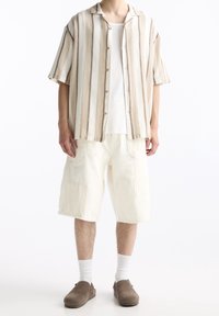 Camicia a maniche corte a righe beige e marrone, abbinata a pantaloni cargo bianchi. Le scarpe slip-on marroni completano l'outfit.