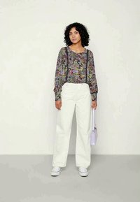 Blusa floral de manga longa com punhos franzidos, combinada com calças largas brancas de cintura alta e ténis brancos; a segurar uma mala lilás clara.