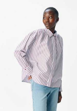 Kvinde iført en hvid skjorte med røde lodrette striber og lyseblå jeans, der ser ud til siden med den ene hånd i lommen.