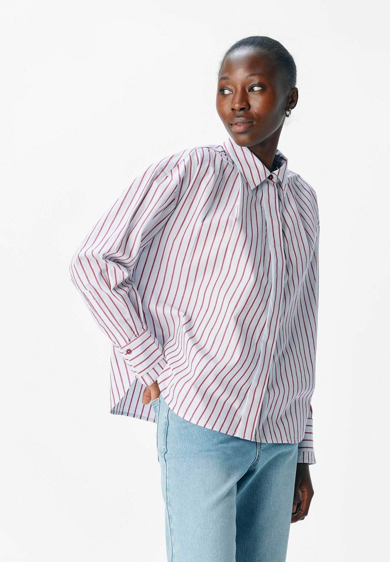Kvinna iklädd en vit skjorta med röda vertikala ränder och ljusblå jeans, som ser åt sidan med en hand i fickan.