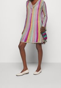 Abito a righe multicolori con scollatura a V e dettagli con nodo, realizzato in un tessuto lucido e texturizzato. Indossato con scarpe flat di colore chiaro e una borsa piccola.