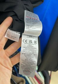 Sort tøjlabel med vaskeanvisninger og mærkeoplysninger. Tags indeholder flere sprog og certificeringssymboler fra agenturer.