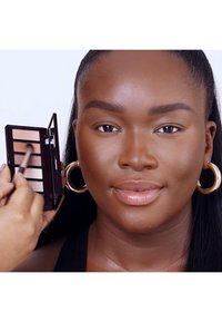 Palette di ombretti con sei tonalità in un astuccio compatto, che presenta una mano che applica il trucco su una pelle liscia di tonalità media, con orecchini a cerchio.