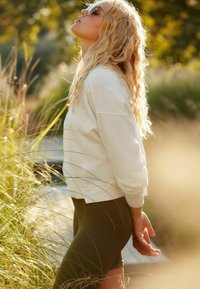 Witte sweatshirt met verlaagde schouders, olijfgroene leggings, blond golvend haar, met een zonnebril op, staand tussen hoog gras in het zonlicht.