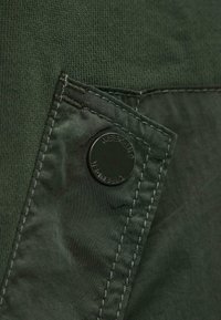 Groene jas met een gestructureerde stof met een gladde afwerking, een zichtbare knop met de opdruk "WEEKEND" en donkere stiksels langs de naden.