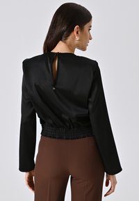 Blusa nera in satin con collo alto, vita elastica e dettaglio a occhiello sul retro. Maniche lunghe e texture liscia con una leggera lucentezza.