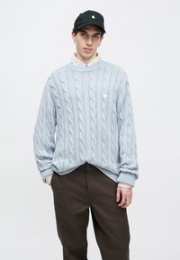 Ανοιχτό μπλε πλεκτό πουλόβερ με στρογγυλή λαιμόκοψη, ριμπ μανσέτες και ρίγα στο τελείωμα. Συνδυασμένο με σκούρο καφέ παντελόνι. Μαύρο καπέλο με λογότυπο.