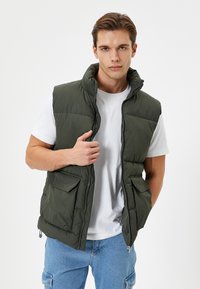 Chaleco acolchado verde con cuello alto, con un cierre frontal y dos grandes bolsillos laterales. Lleva puesto encima de una camiseta blanca y pantalones de mezclilla azul.