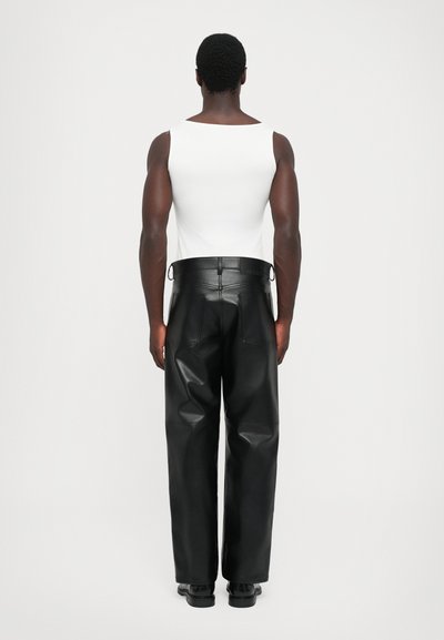 Homme de dos portant un haut sans manches blanc et un pantalon large en cuir noir taille haute avec des chaussures noires sur fond uni.