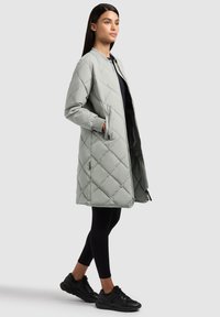 Naine seisab, kandes pika, helehallist tikandiga mantlit mustade retuuside ja mustade tossudega, käed mantlitaskutes, vaade kergelt paremale.