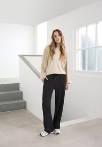 Haut beige à manches longues avec des manches côtelées, associé à un pantalon noir à jambes larges et des baskets grises, se tient sur un sol clair près d'un escalier.