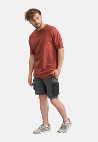 Chemise rouge à manches courtes avec une coupe décontractée, portée avec un short cargo gris comportant des poches latérales et une touche de teal. Des chaussures de sport grises complètent le look.