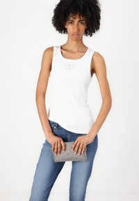 Weißes geripptes Tanktop mit dezentem Logo, kombiniert mit blauen Jeans. Hält eine strukturierten grauen Clutch mit metallischem Glanz.