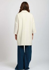 Cappotto crema oversize con maniche a tre quarti, realizzato in tessuto morbido, con taglio dritto e design minimalista, abbinato a pantaloni in denim a gamba larga.