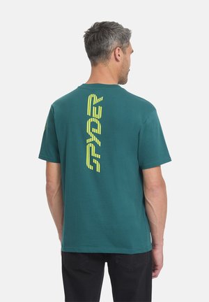 Mann trägt ein türkisfarbenes Kurzarmshirt mit vertikalem gelben "PYRE" Schriftzug auf dem Rücken, kombiniert mit schwarzer Hose, steht und blickt nach rechts.