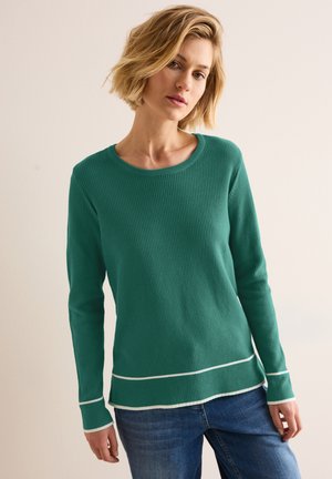 Chemise à manches longues verte côtelée avec un col rond et une bordure blanche à l'ourlet et aux poignets, assortie à un jean en denim bleu.