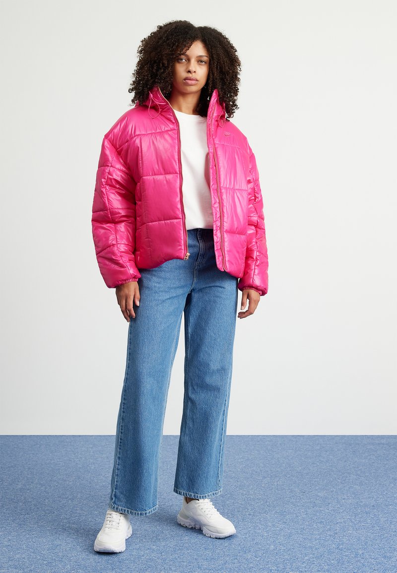 Rosa Puffjacke mit einem glänzenden Finish, ausgestattet mit einem hohen Kragen, Reißverschluss und quadratischem Steppmuster, kombiniert mit hellblauen Jeans und weißen Sneakers.