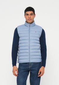 Világoskék bélelt mellény magas gallérral, cipzáras záródással és oldalzsebekkel, tengerészkék hosszú ujjú ing fölött viselve. Klasszikus logó a mellkason.