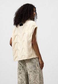 Jasno kremowy, dziany kamizelka z falistym wzorem i krótkimi rękawami, w zestawieniu z szarymi, marmurkowymi spodniami o szerokich nogawkach. Prezentuje swobodny, teksturowany wygląd.