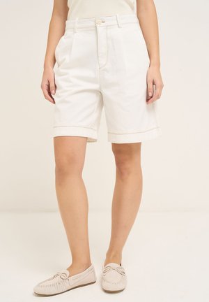 Kvinde iført high waist, off-white knæshorts kombineret med lysebeige mokassinsko, stående mod en ensfarvet baggrund.
