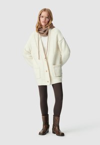 Cardigan bianco lavorato a maglia con tasche frontali e bottoni marroni, indossato sopra una maglietta chiara. I leggings marroni e gli stivaletti alla caviglia marroni completano l'outfit.